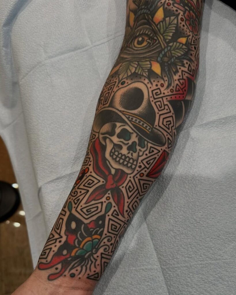 A forearm featuring various American traditional style tattoos including a skull and geometric patterns.