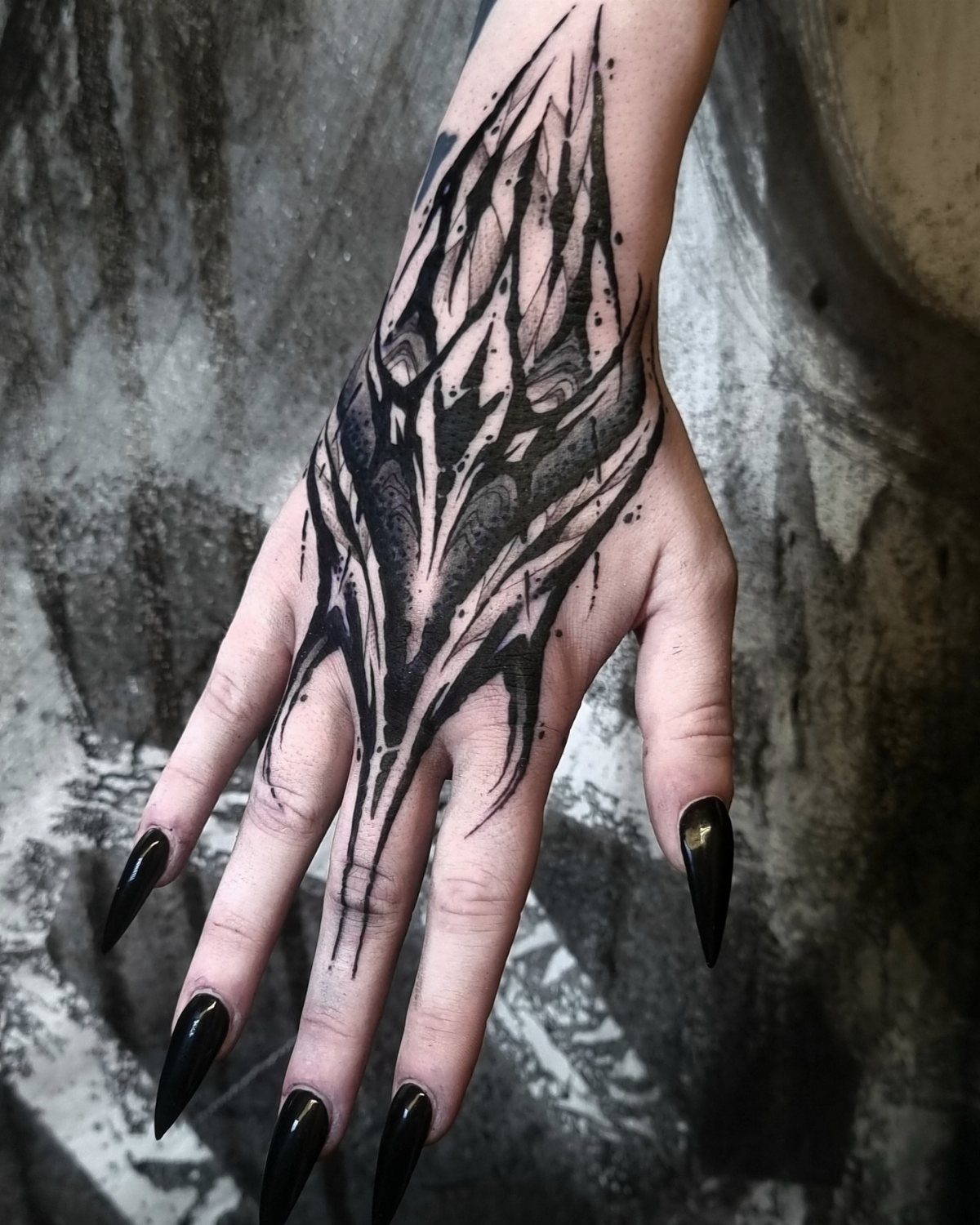 A hand with a sharp blackwork tattoo and long black stiletto nails against a textured background.