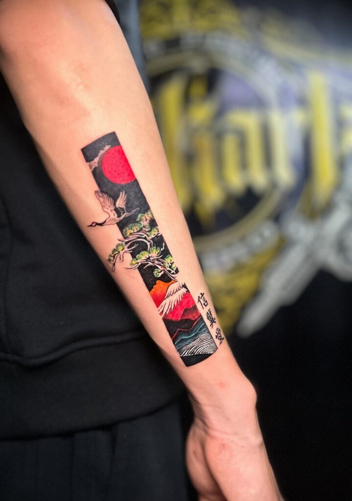 A colorful Japanese style forearm tattoo featuring cranes, a red sun, and mountains.