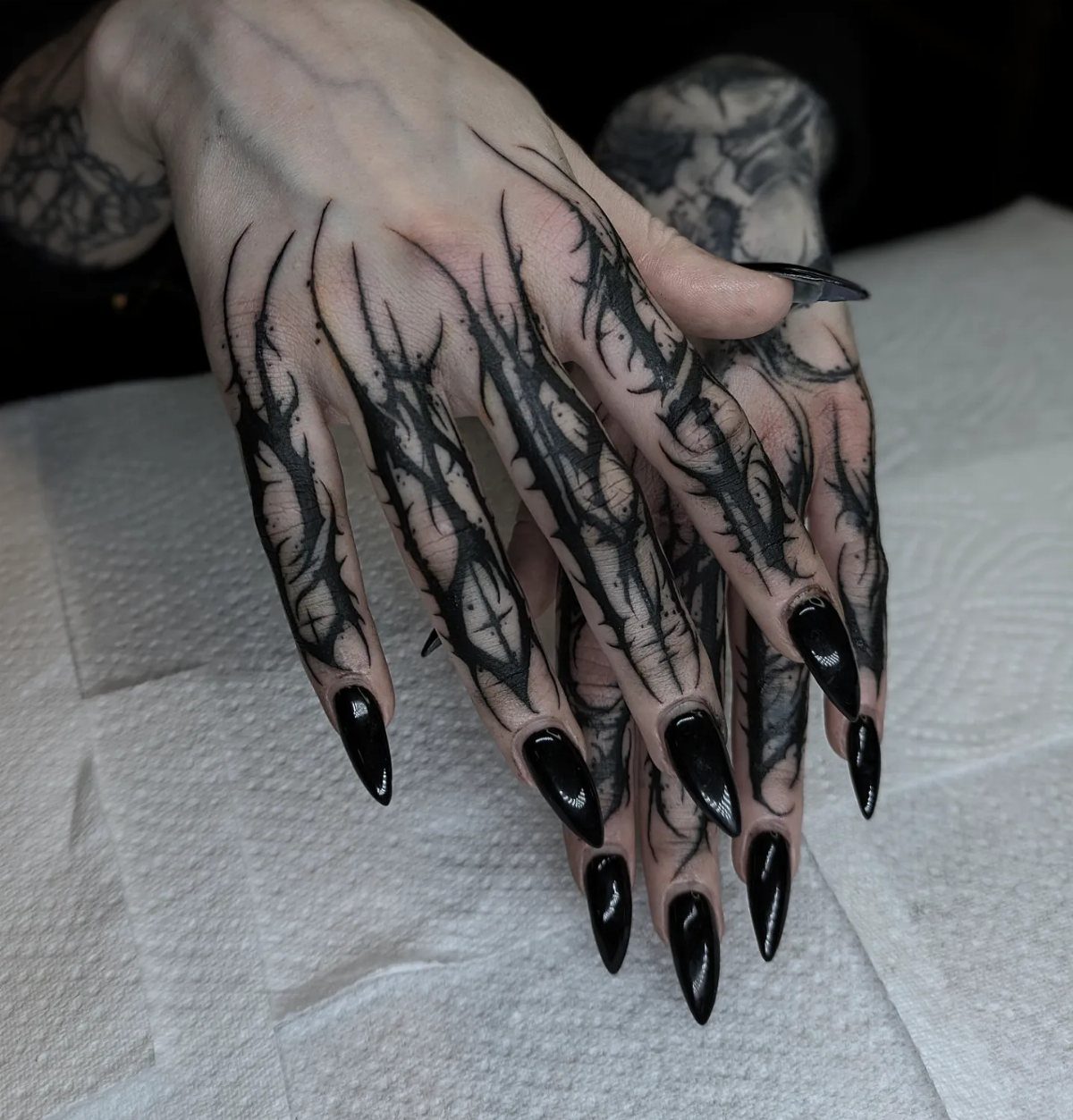 Hands with black thorny finger tattoos and long pointed black stiletto nails on white paper.