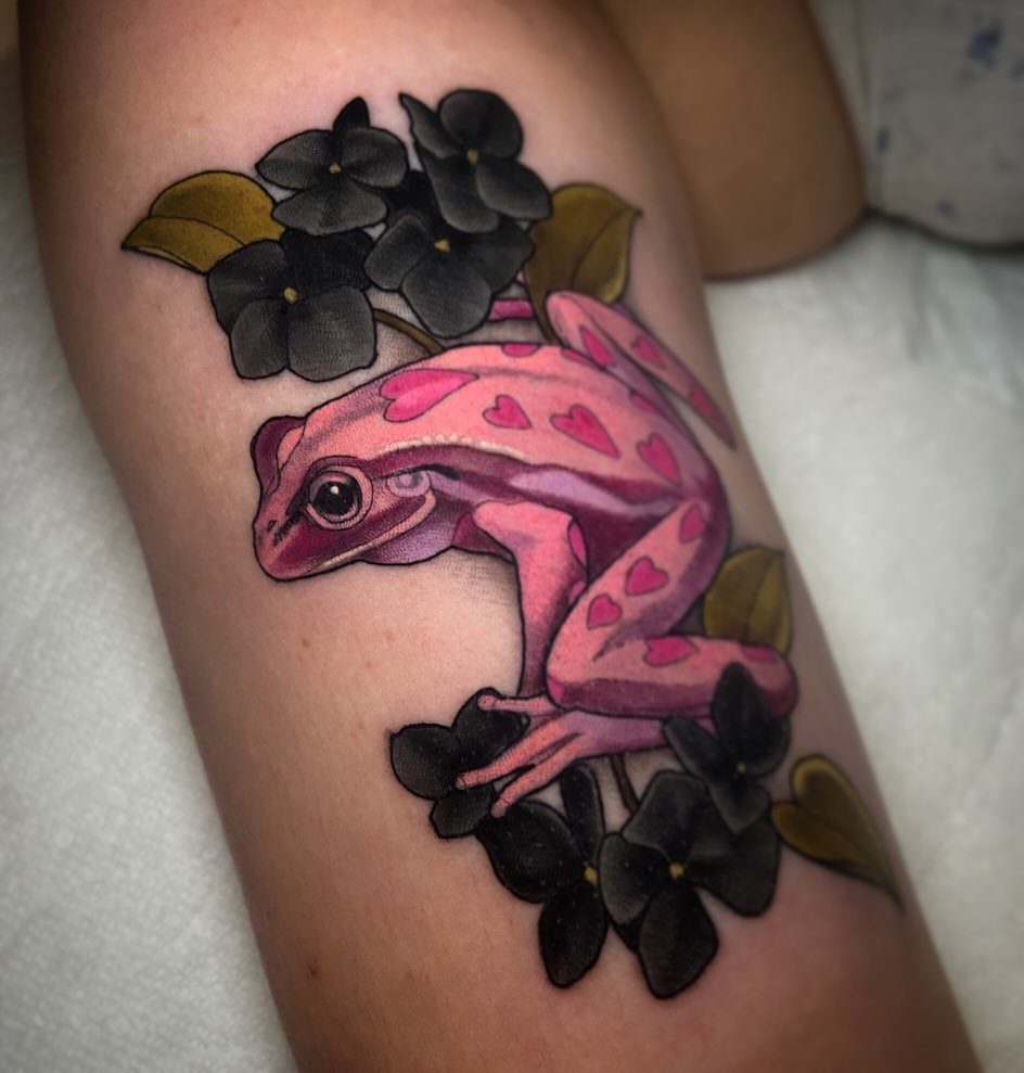 A pink frog tattoo with heart patterns surrounded by black flowers on human skin.
