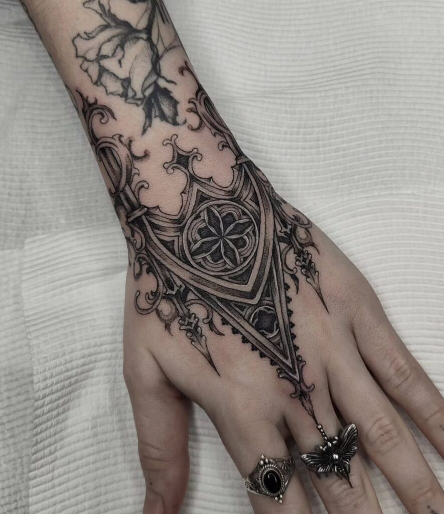 A hand and forearm decorated with intricate black ink gothic style tattoos and dark rings.