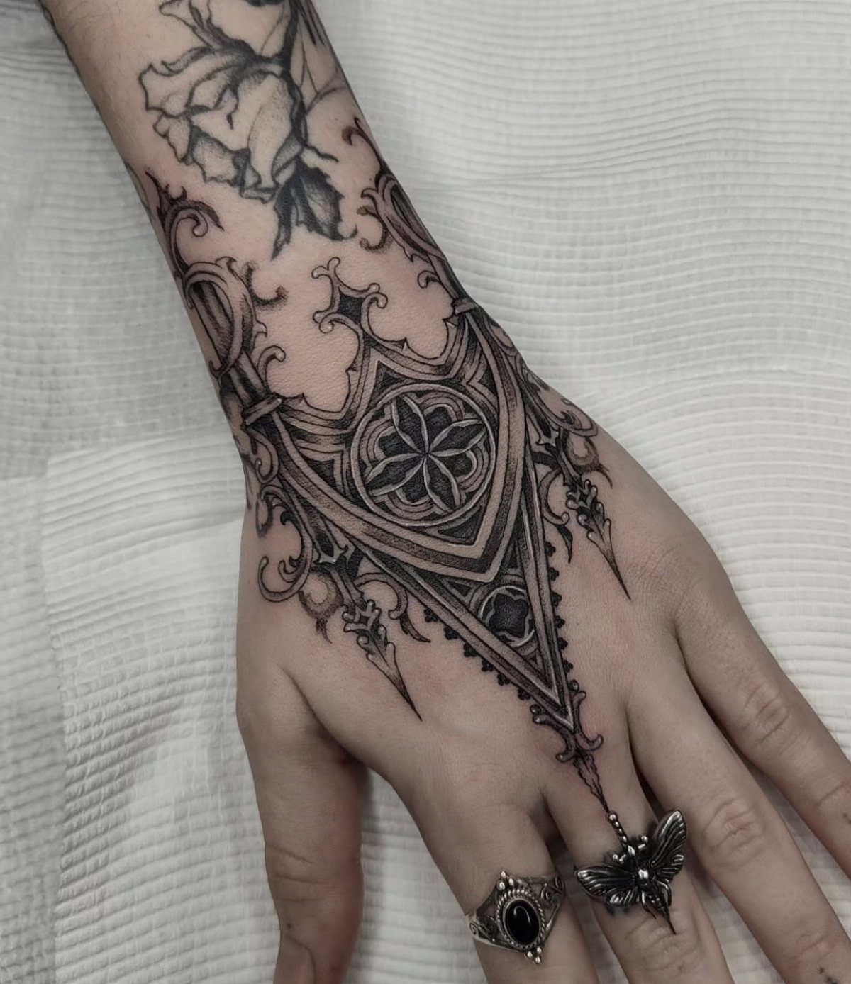 A hand and forearm decorated with intricate black ink gothic style tattoos and dark rings.