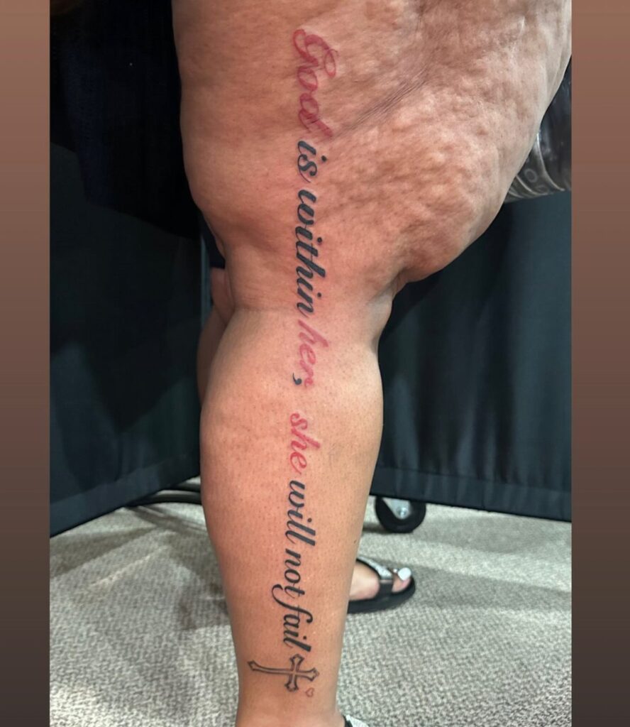 A vertical script tattoo on a person's leg reading God is within her she will not fail.