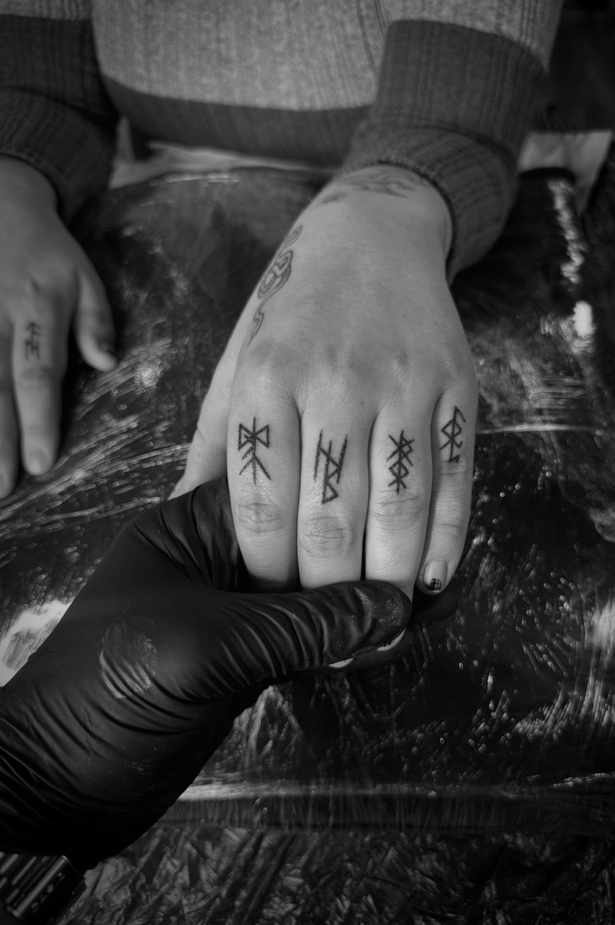 A hand with small rune tattoos on the fingers being held by a gloved hand.