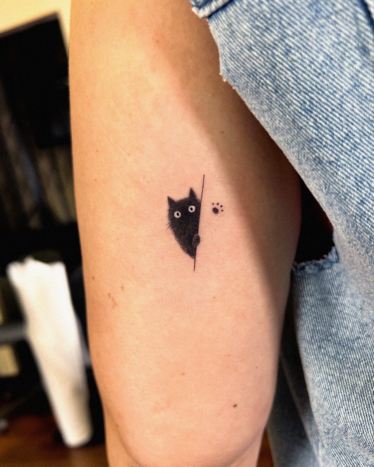A small minimalist black cat tattoo peeking from behind a line on a person's arm.