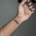 A small black cursive tattoo reading embrace on a person's inner wrist.
