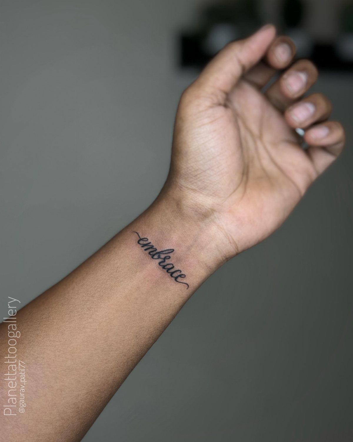 A small black cursive tattoo reading embrace on a person's inner wrist.