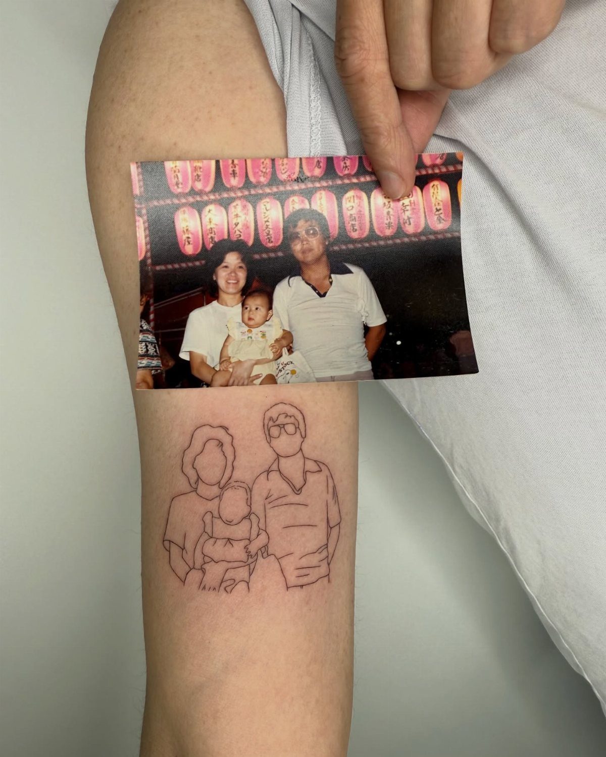 A hand holds a family photograph above a matching fine line outline tattoo on an arm.