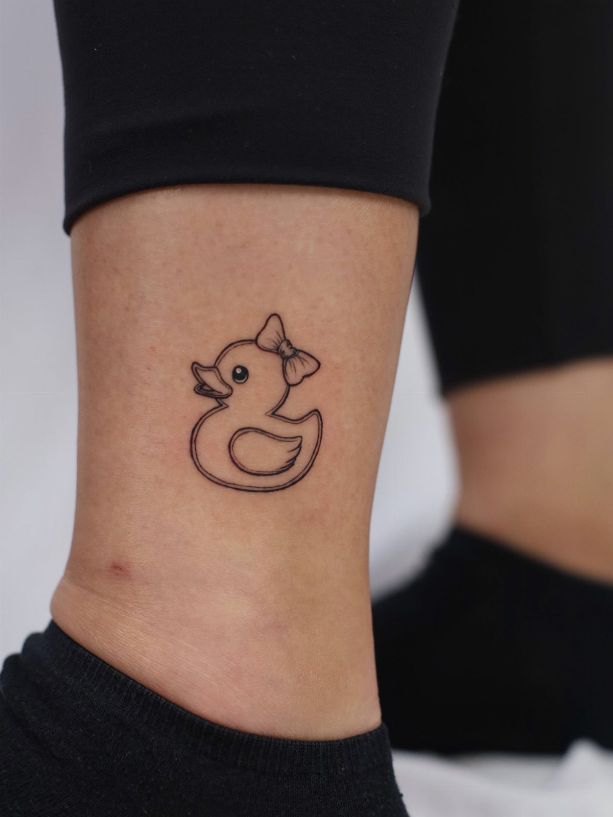 A small black ink tattoo of a rubber duck on a person's ankle.
