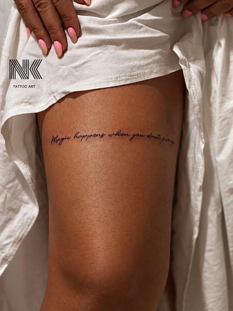 A script tattoo reading 'Magic happens when you don't give up' on a person's thigh.