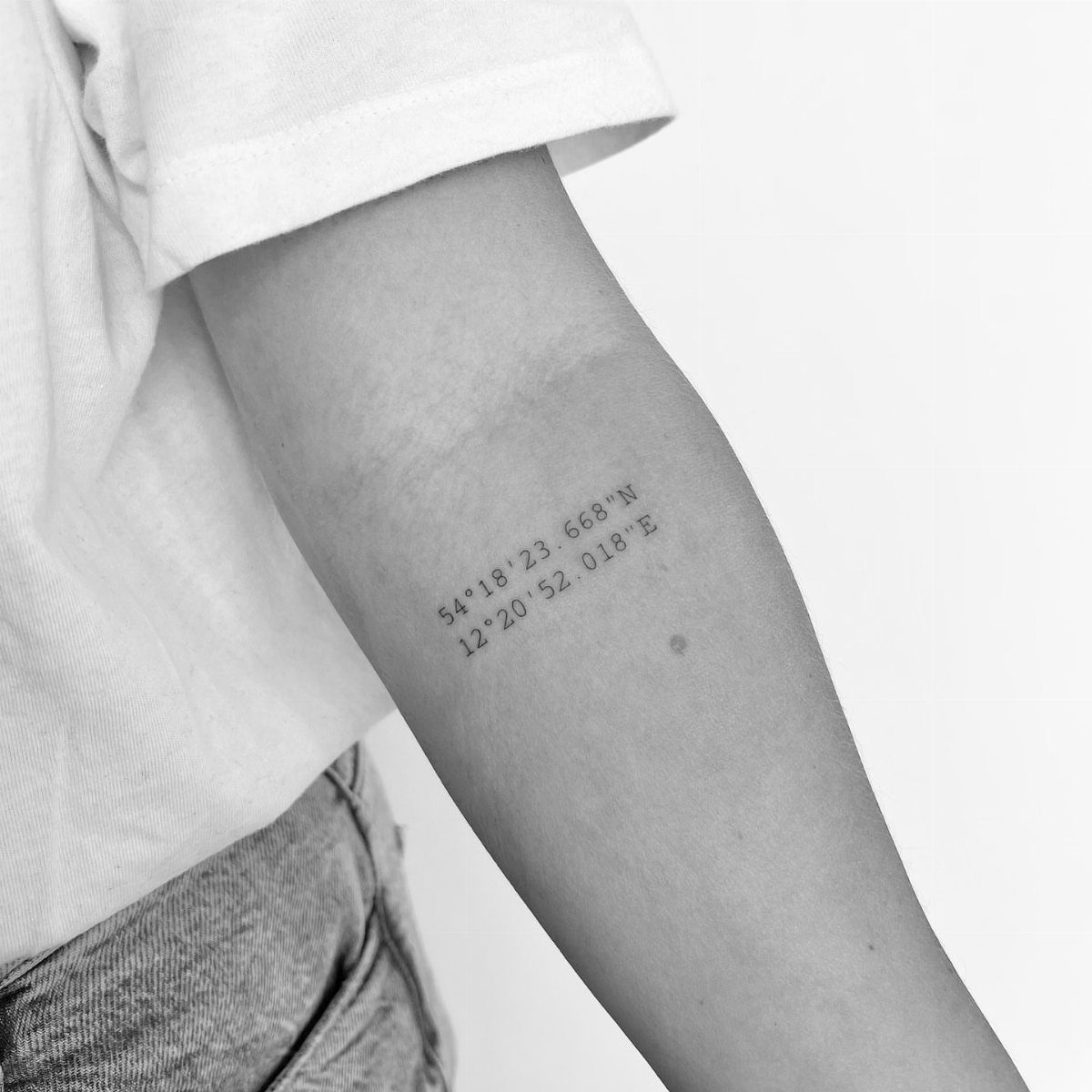 A small tattoo of geographic coordinates on a person's forearm in black and white.