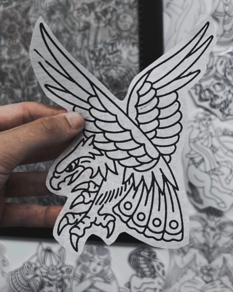 A hand holding a paper stencil of a traditional black line art eagle design.