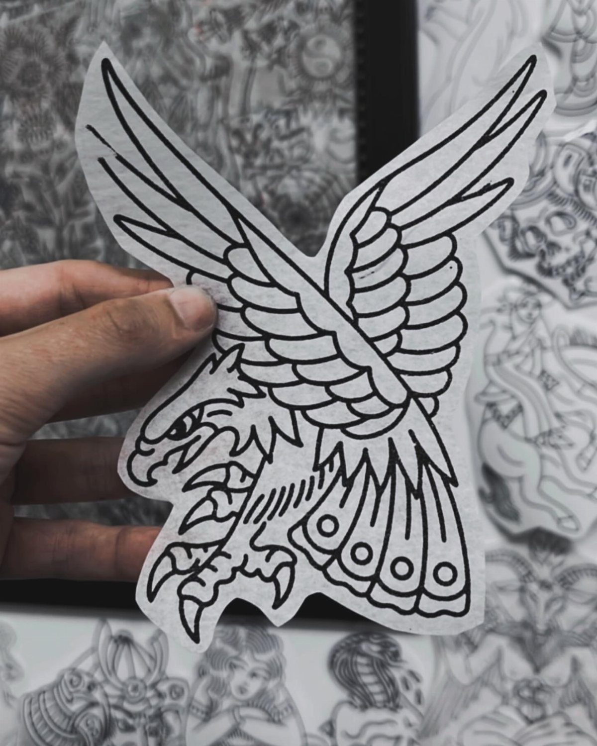 A hand holding a paper stencil of a traditional black line art eagle design.