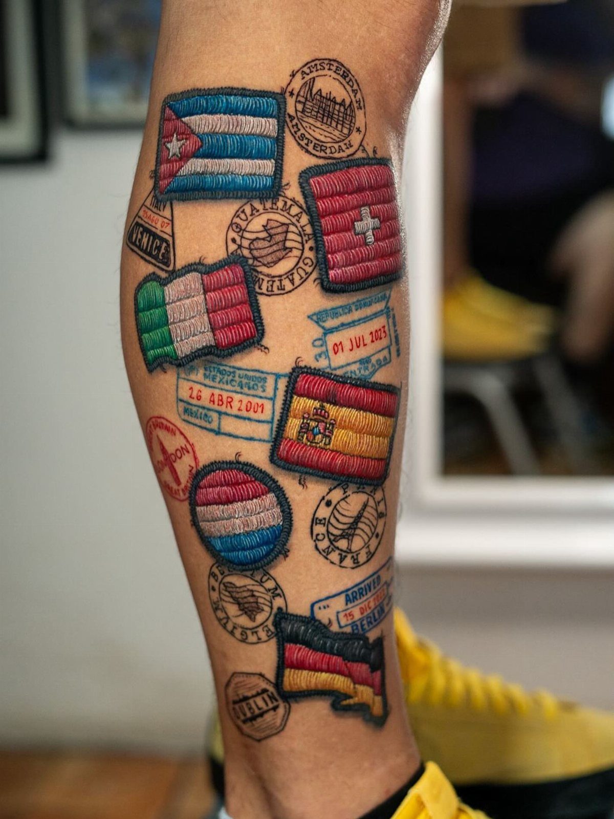 A person's leg covered in colorful tattoos designed to look like embroidered travel patches and flags.