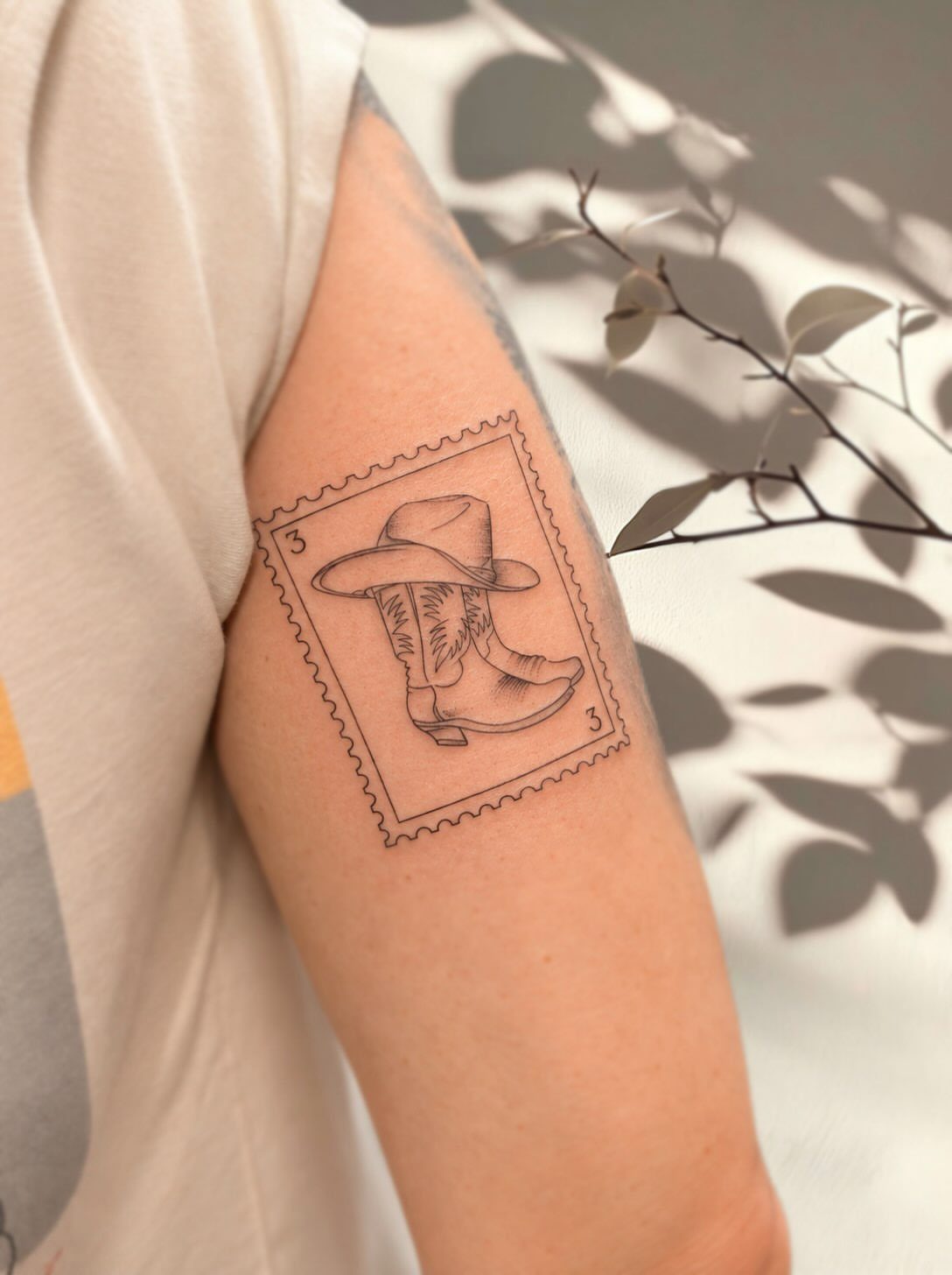 A fine line tattoo of a cowboy hat and boot inside a postage stamp border on an arm.